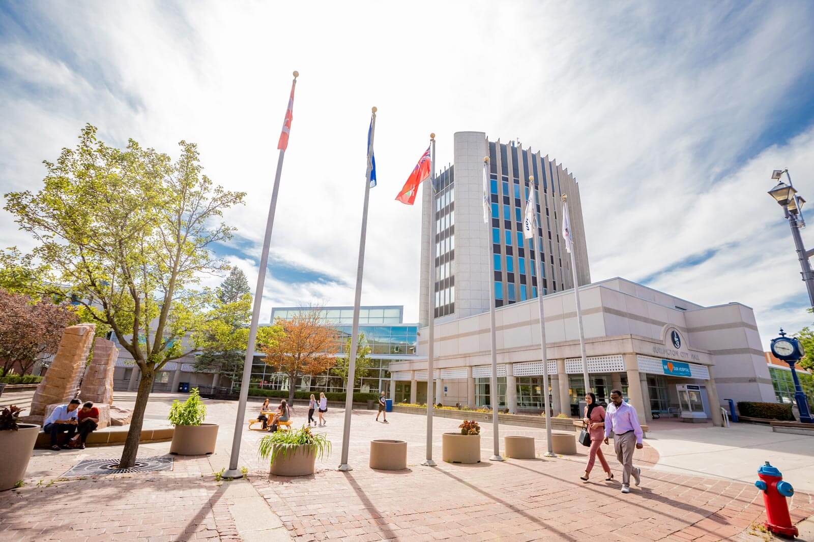 Image Description: A photograph of the Burlington Civic Square on a sunny day. Pedestrians walk through the open area with flag poles, plants, and benches. The city hall building is in the background
